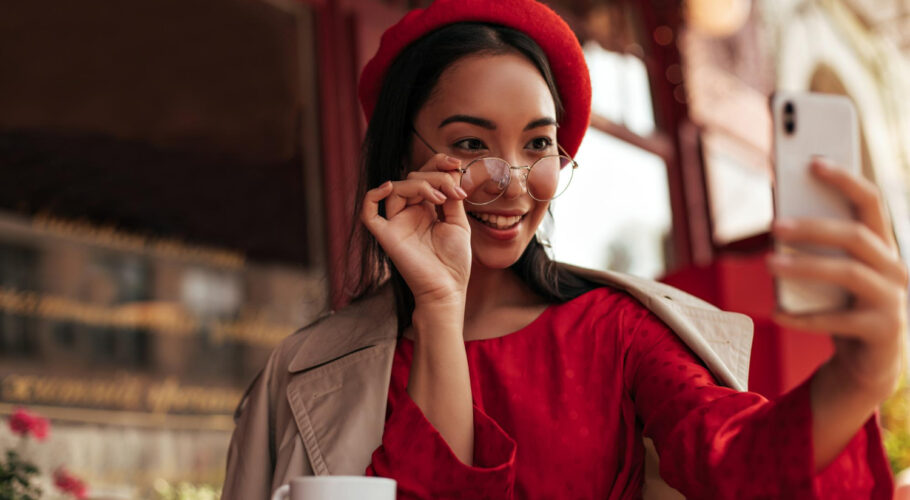 attractive brunette woman trendy beret red dress beige trench coat puts off eyeglasses holds phone takes selfie street cafe china