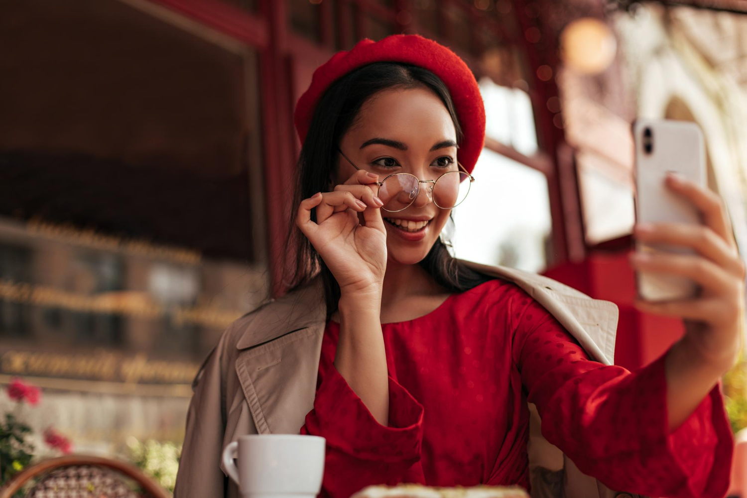 attractive brunette woman trendy beret red dress beige trench coat puts off eyeglasses holds phone takes selfie street cafe china