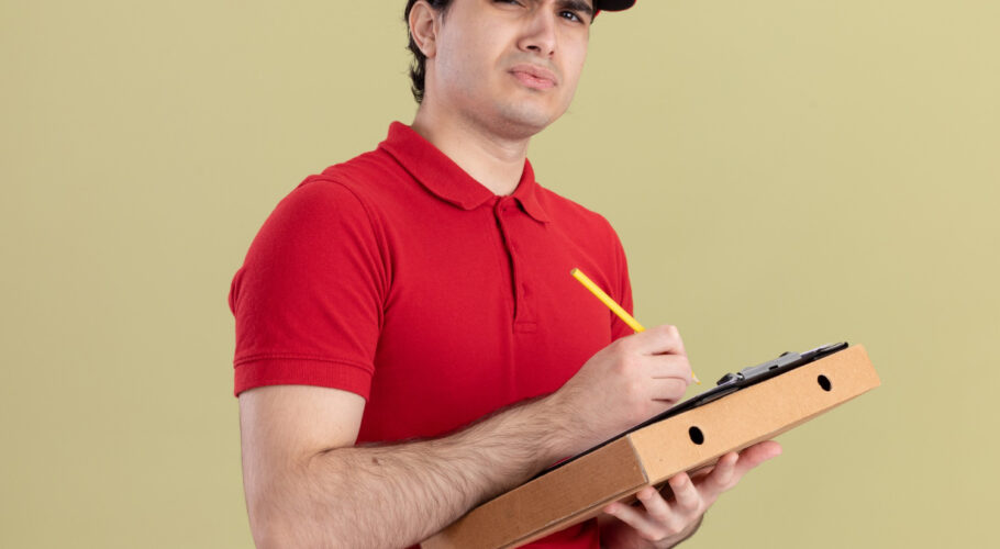 confused young caucasian pizza hut delivery man in red uniform holding pizza and clipboard
