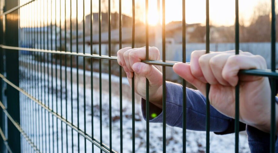 hands hold onto metal fence of a prison with sun in background