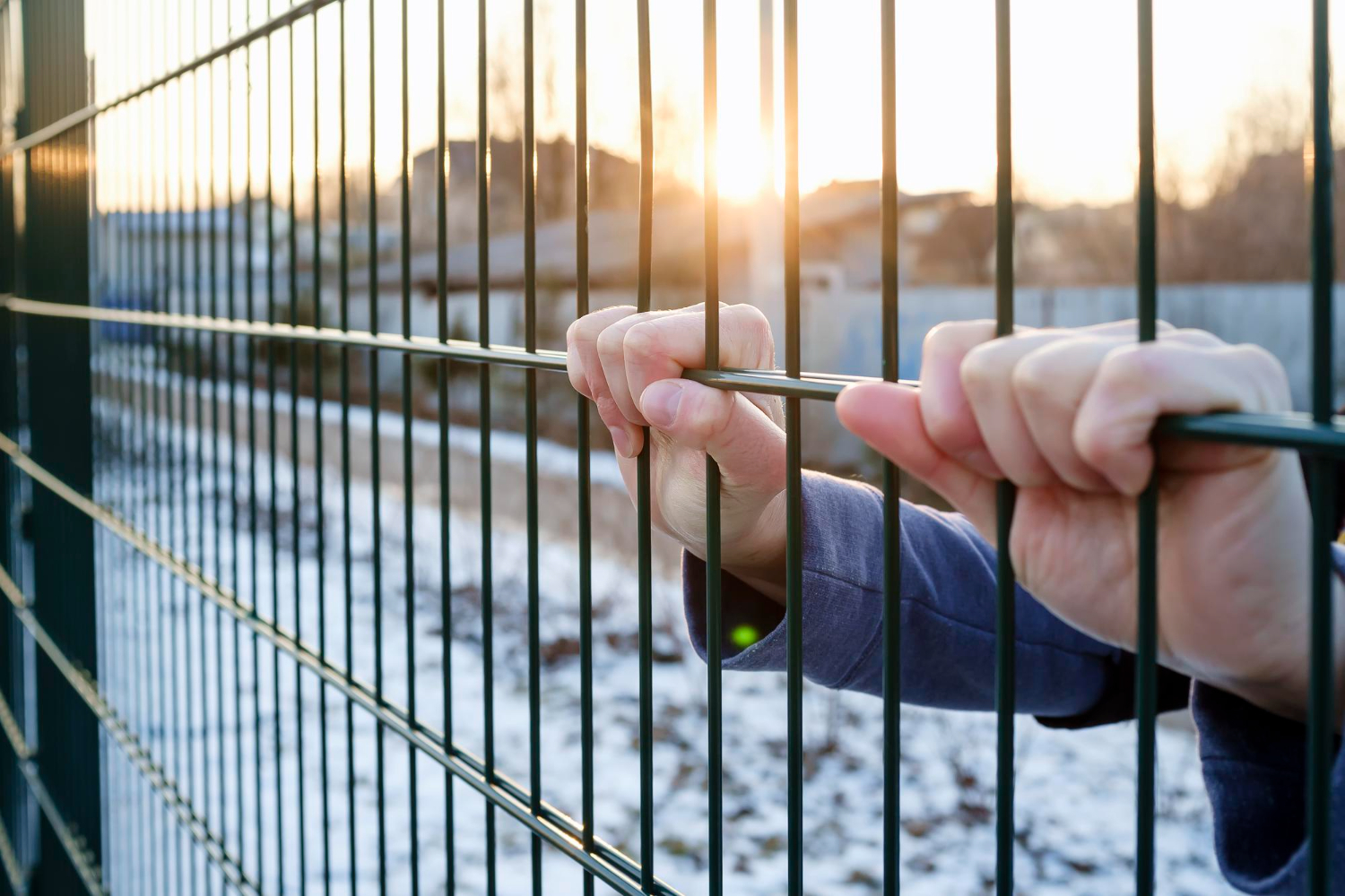 hands hold onto metal fence of a prison with sun in background