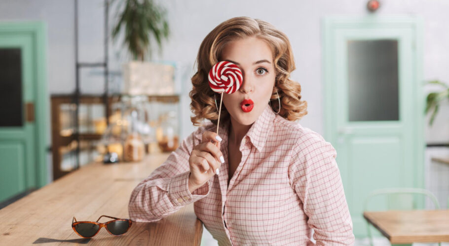 pretty lady sitting bar counter covering her eye with lollipop candy while amazedly looking at camera in 1950s cafe