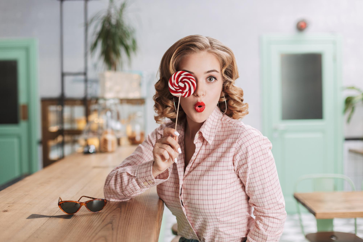 pretty lady sitting bar counter covering her eye with lollipop candy while amazedly looking at camera in 1950s cafe