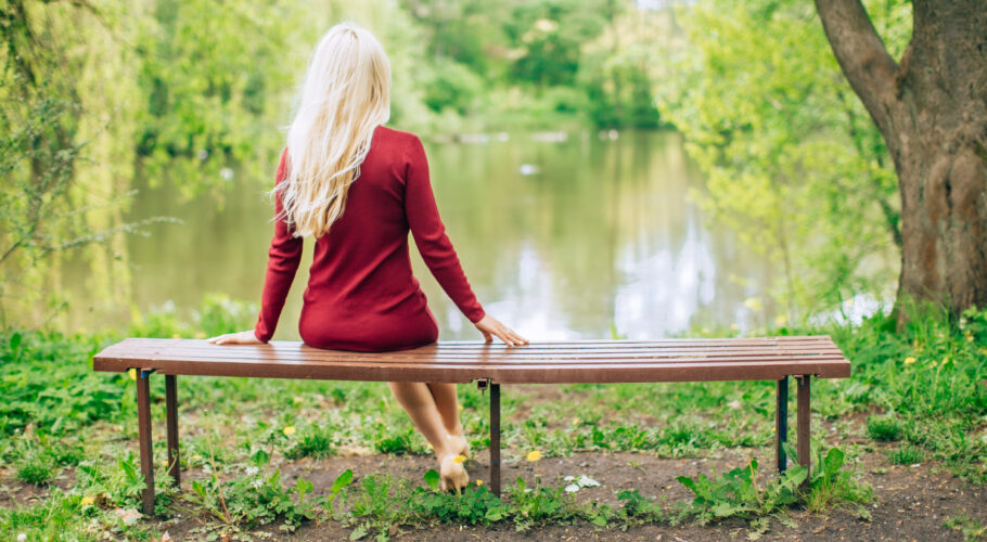 young blonde woman jewell red dress sitting bench park with lake view