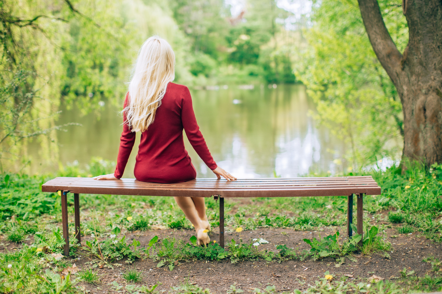 young blonde woman jewell red dress sitting bench park with lake view