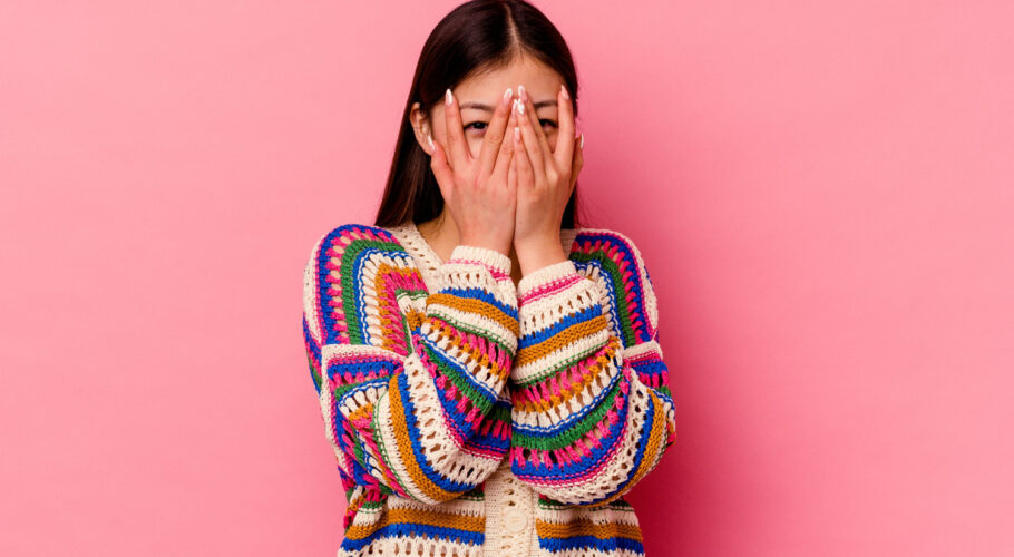 young chinese woman isolated pink wall blink through fingers embarrassed covering face