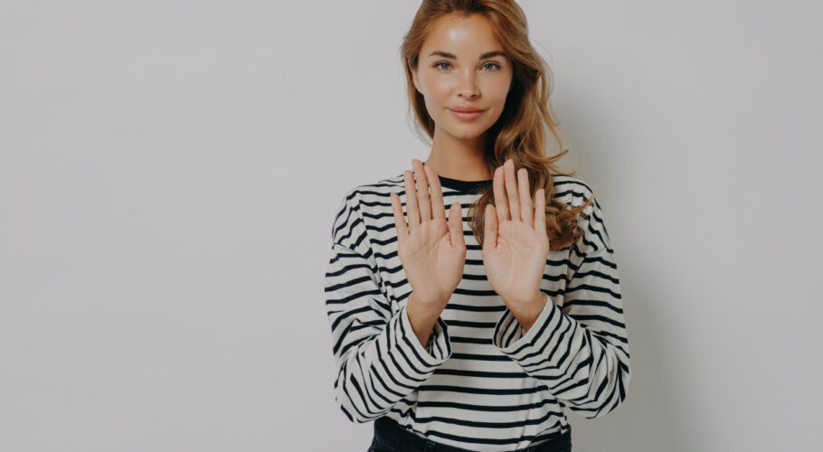 young confident woman gesturing stop sign saying no while standing isolated against grey wall