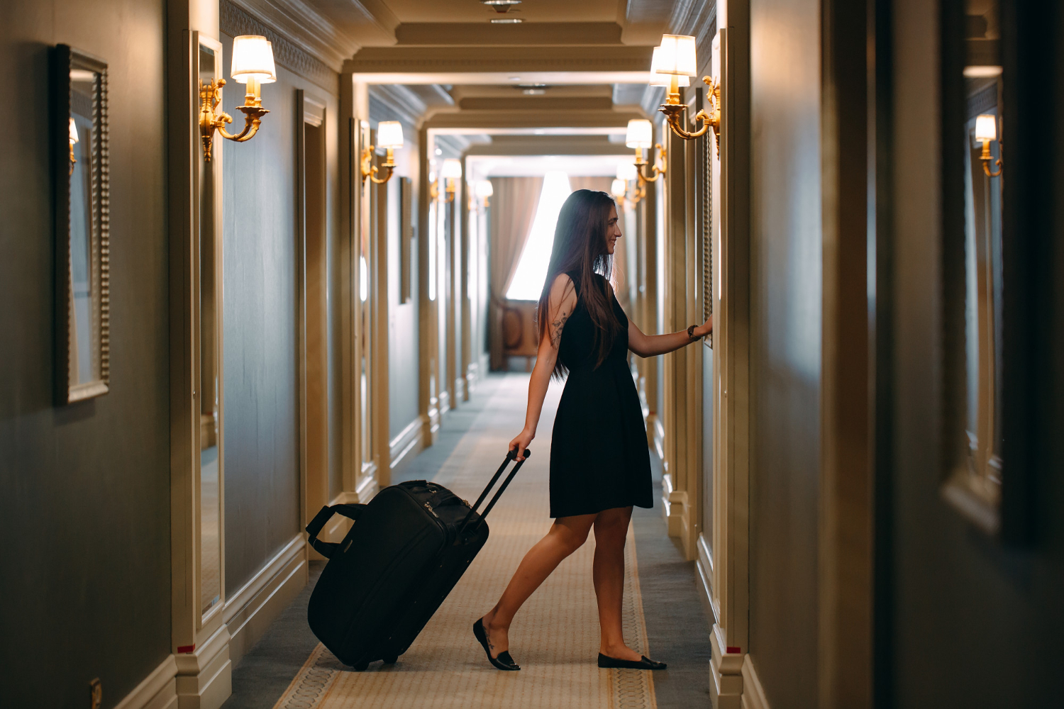 young elegant woman with handbag and suitcase walks down hotel corridor to her room